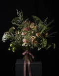 A rustic dried flower bouquet with green foliage, beige and pink wildflowers, and a dusty rose ribbon, displayed on a dark pedestal against a black background.