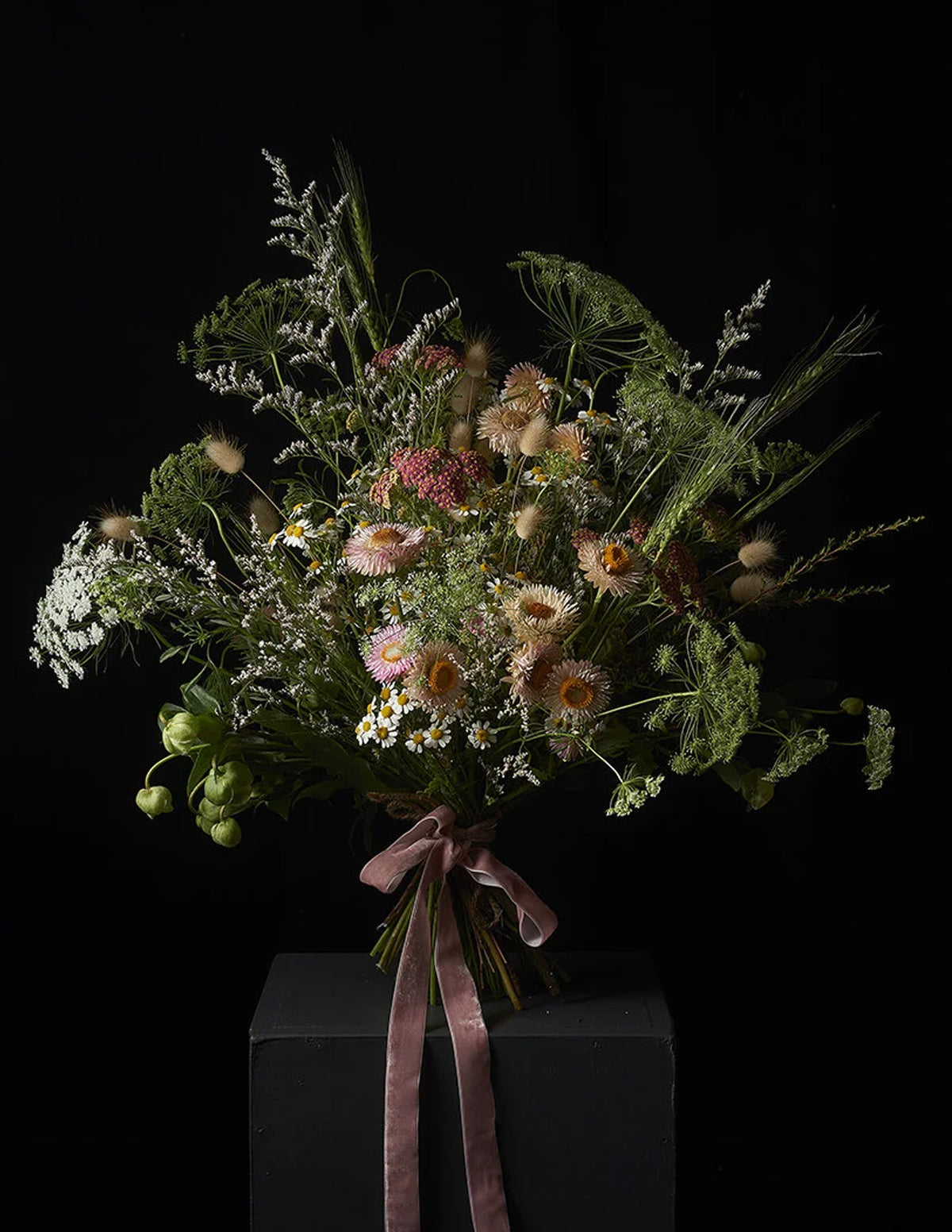 A rustic dried flower bouquet with green foliage, beige and pink wildflowers, and a dusty rose ribbon, displayed on a dark pedestal against a black background.