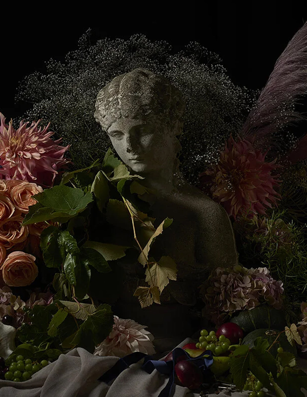 A dark, moody still life featuring a classical stone bust surrounded by abundant flowers, green leaves, grapes, and soft fabric against a black background.